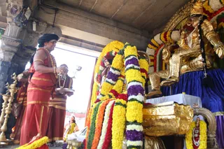 A photograph of a temple shrine with a devotee in a red and gold dress standing at left holding a silver puja vessel and a small silver bowl, with a black turban and a long beaded necklace. Behind them a second person in white and gold stands near a pillar. At right a large golden deity murti seated on a blue velvet-draped pedestal is decorated with thick garlands of yellow marigolds, purple and white flowers, and red and orange blooms. A golden ornamented stand and brass lamps sit in the foreground.