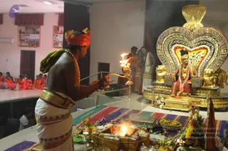 A homa ritual in an indoor hall: a priest in white dhoti with a bare chest, an orange head cloth and a green shawl over his shoulder holds a lit brass arati lamp over a fire pit of wooden logs and offerings, with a bowl of flowers and fruit nearby. Behind him stands an ornate golden throne with a cobra-hood backrest decorated with coloured beads and flowers, where a figure in orange and red garments with a crown and a garland sits. Several people in saffron robes stand near the throne, and a group of devotees in orange sits on a stage at left. Two framed posters hang on the wall. Colourful striped mats cover the floor.