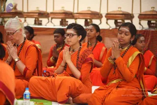 A photograph of a group of women seated on the floor in orange and saffron robes during a ritual, hands pressed together in namaste. Several wear rudraksha malas and white vibhuti lines on their foreheads. The front-left woman has white hair and glasses; the central woman wears glasses and a red-bordered orange shawl. Behind them, brass covered vessels line a raised platform, and water bottles and small offerings sit on the floor.