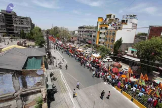 Elevated view of a wide urban street in an Indian town during a daytime procession. A long line of people walks along the road, many in white and light clothing, alongside decorated carts and open vehicles draped with orange, red and white cloth and canopies. Saffron and orange flags and banners fly from poles along the route. Parked white cars, two-wheelers, multi-storey buildings with balconies and a few pedestrians on the pavements surround the procession. Trees and a blue sky frame the scene. The number of people visible is large, with dense crowds along the procession line.
