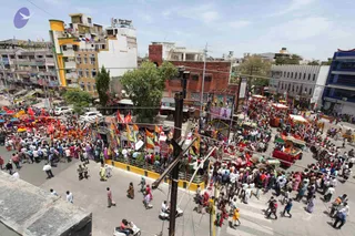 Aerial view of a dense street procession in an Indian town, with a large crowd of people filling the road and pavements. Many people wear white, pink and bright clothing, and many carry or wave red, orange and yellow flags and banners. Decorated carts and float-like vehicles with canopies and red draped sections move through the crowd near a multi-storey building with colourful facades, and a small pavilion stands at the centre. Scooters and parked cars line the road, and utility poles and overhead wires cross the scene.