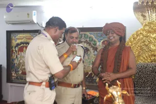 A photograph of an indoor room with a gold-carved throne-like chair at right and framed traditional painted panels on the wall. Two uniformed police officers stand at left: one in a light khaki shirt with a radio at his belt, the other in khaki with a brown leather belt, holding a glass of a light drink and looking at a mobile phone. At right, a smiling person in an orange-saffron robe and an orange turban, wearing a rudraksha mala and a bead necklace, stands with hands clasped beside a small gold ritual object. A leopard-print cushion and an air conditioner are visible. a person: the person in saffron with turban and rudraksha mala is a candidate by appearance, but the cues are limited, so the identity is uncertain.