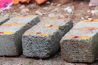 Close-up photograph of several grey concrete bricks laid on bare reddish-brown earth with small stones and grass. Each brick top is marked with a bright yellow turmeric band and a red kumkum dot. Pink and orange fabric is visible at the top edge of the frame. No people are visible.