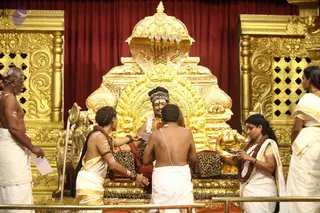 Photograph of a darshan scene on an ornate gold throne (peetha) with a red curtain behind it. A seated figure in red-orange clothing with a striped headwrap and garland sits on a leopard-print cushion, while devotees in white dhotis and saffron-bordered white sarees approach to offer and receive blessings. One man in a white dhoti with a sacred thread stands bare-chested in front of the throne, and a bearded older man in white with forehead markings stands at the left edge. A woman in a white saree with rudraksha mala stands at right, and another woman in a cream saree stands at the far right. Gold lion-motif armrests and carved gold panels frame the dais. Six people are visible, the seated figure included.