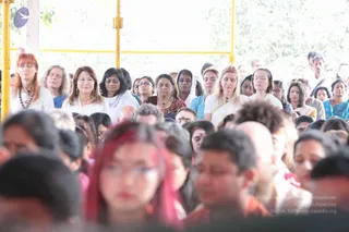 A crowd of devotees sits closely together under a covered open-air pavilion with yellow metal poles, eyes closed in meditation or prayer. Foreground figures include a woman with bright red hair and glasses in a dark top, and a bald man in a saffron-toned shirt. Behind them, women wear white kurta-style clothing with shawls, one woman in red and orange patterned dress, others in blue and teal saris and salwar, some with bindi and red tilak marks. Men in white stand at the back. The scene conveys a large satsang or darshan gathering, with many people visible. No a person candidate is clearly identifiable in this frame.