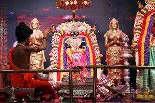 Photograph of a temple puja for a deity murti seated on an ornate throne with a gold-toned canopy and a red, purple and yellow flower garland arch. Two gold-coloured attendant figures with hands in anjali stand on either side. A man seated at left, bare-chested with long dark hair piled up, wears an orange saffron cloth, multiple rudraksha-style beaded necklaces and bracelets, and raises his hands in ritual gesture toward the deity. Silver and brass puja vessels, a lamp, pink flowers and a lectern are arranged in front. A sari-clad figure in green and pink stands at right. Dark stage backdrop with a starry projection.