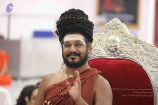 A man seated on an ornate silver throne with a red velvet seat and a carved sun-face motif gestures with his right hand raised. He wears a rust-orange shawl with rudraksha beads, a gold pendant on a gold chain, and a tall braided dark topknot wrapped with rudraksha beads. White vibhuti marks his forehead. Behind him a blurred hall with an orange-robed figure and a framed picture is visible. a person is shown in a seated discourse setting; his distinctive topknot, rudraksha, gold necklace and orange shawl are the qualifying cues.