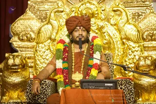 A seated man in a rust-orange turban with a crescent emblem and a red bindi sits on an ornate gold throne carved with lion heads and scrolled panels, flanked by golden lion statues on leopard-print cushions. He wears an orange robe, layered gold necklaces and a long mala of red, yellow, green and white flower garlands, with a microphone before him and a Yamaha mixer on a draped orange table. Swamiji (probable) is shown in this satsang setting; the appearance cues are the turban, orange robe, garlands and ceremonial gold necklace.