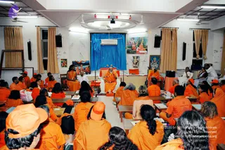 A seated satsang in a plain room with white walls, ceiling fans, fluorescent lights and beige curtains. A bearded man in a saffron-orange robe sits centrally on a raised orange-draped dais with microphones, flanked by a white-clad attendant. Behind him hang a blue backdrop and framed religious pictures. Rows of listeners in saffron-orange clothing and orange caps sit on wooden benches, some with books. A woman in orange stands at left near the dais and another seated figure in orange sits at right. The central man has long hair, a crown-like adornment, and a rudraksha-style necklace with saffron clothing, a person candidate.