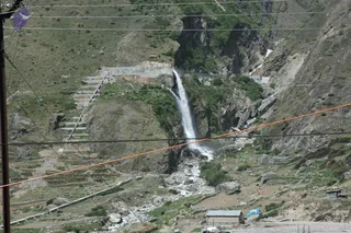 A photograph of a mountain landscape with a waterfall cascading down a steep rocky gorge in the Himalayas. Stone steps climb a terraced green slope on the left, and a concrete channel runs along the cliff above the falls. A stream flows through rocks at the bottom, beside a small stone-walled hut with a metal roof and a blue tarpaulin. Overhead power lines and a utility pole cross the scene. No people are visible.