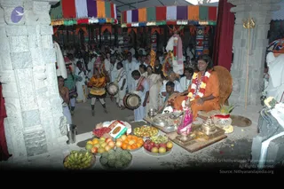 A photograph of a temple courtyard during a ritual. A seated figure in saffron-orange robes and a red-and-white flower garland sits on a carved wooden chair with a shaped backrest, behind a low table laden with bananas, oranges, apples, pomegranates and other fruit, plus a brass lamp, metal bowls and a pink-wrapped object. Around them, people in white clothes and dhotis stand and sit beneath tall pillars and a striped cloth canopy in purple, orange, green and white. A drummer in a yellow costume with a tall headdress plays a drum, a performer in red and green stands nearby, and a golden trident is mounted on a pillar at right. Several people wear white, and the setting appears to be a festive temple ceremony.