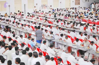 A large indoor hall filled with many men and women in white clothes seated at long grey tables with red plastic chairs, eating from plates of food. Some attendees in white dhotis and shirts stand between the rows, and a server in a striped shirt moves through the aisle. At the far back a figure in white stands near a framed picture on the wall. The scene shows a community meal, annadan-style, during a multi-day programme.