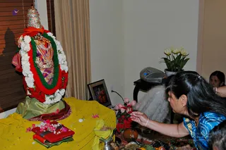 A woman in a blue patterned sari with gold border reaches toward a small altar holding a decorated deity murti. The murti wears a tall jeweled crown, a red and gold garment, a green and red garland and white flower garlands. In front are pink and red roses, yellow cloth, a red-covered book, fruit, a small brass vessel and a framed portrait of a man in red robes on a side table. A white potted tulip-like plant sits nearby, and another woman is partly visible at the right edge. No a person candidate is clearly visible.