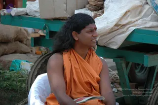 Outdoor photo of a man with long, swept-back dark hair seated on a white cloth beside a teal-painted cart loaded with cardboard boxes and sacks. He wears a saffron-orange draped shawl over his shoulders and a red tilak mark on his forehead, and holds a small open booklet in his lap. a person: long hair, saffron draped clothing and a tilak mark, seated in the cart area during the Kodikkan ratham journey back to Bidadi.