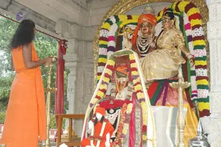 A devotee in an orange sannyasi-style robe stands at left, holding a brass aarti lamp or offering vessel toward a shrine. Inside a stone pavilion with a gold-framed arch, two large deity murtis are dressed in gold and draped in layered garlands of yellow, red, white and green flowers. The seated figure wears a gold saree and gold jewellery, while the standing figure wears an orange turban and a pearl necklace. Brass oil lamps stand on both sides, and a small statue in orange with white flower garlands sits in front of the central pedestal. A white cloth and offerings rest on the pedestal, with trees behind the pavilion.