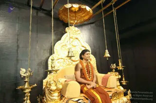 A seated figure sits on an ornate gold throne with a carved gold back and gold lion-head armrests, under a gold canopy hung from the ceiling, with several brass oil lamps and hanging bells in a dark-walled room. Lit oil lamps and bells flank the throne, and a striped tiger-skin style cloth lies on the seat.