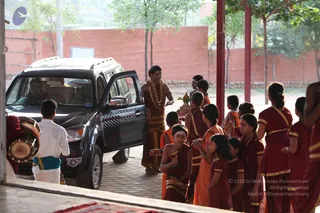 A photograph of a courtyard at an ashram where a dark SUV has stopped under a covered walkway. A young man in a rust-brown dhoti and shawl, with a long rudraksha-style garland and gold-coloured trim, stands beside the open rear door holding a small offering tray. Around him, children and young women in maroon and orange robes with gold borders stand in a line with palms pressed together, and one girl holds a brass plate. A drum rests on the left edge, a man in white dhoti stands near it, and a red wall and green trees form the background. The driver is visible through the windshield. No person is clearly identifiable as a person from this view.