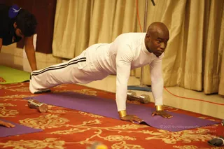 A man in a white long-sleeved top and white track pants with black side stripes performs a push-up on a purple yoga mat over a red carpet with gold floral patterns. He wears gold cuff bands on both wrists and has a shaved head. Two silver dumbbells lie on the carpet beside the mat. Gold curtains hang in the background, and another person in dark clothing is partly visible at the left edge of the frame.