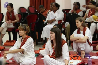 A photograph of a seated congregation during a satsang at an ashram hall with red curtains and red-cushioned chairs. Several women in white ritual clothes wear red-orange shawls and rudraksha-style beaded necklaces. Foreground women sit on red mats with a closed book and water bottles nearby; others on chairs in the background listen attentively.