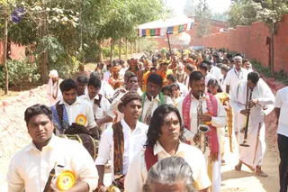 A procession moves along a dusty path between a red brick wall and green trees. In the foreground, men in white shirts and white dhotis play wind instruments: a long silver trumpet-style horn, a wooden clarinet-like pipe and a drum slung on a strap. Several wear orange-and-yellow rosette badges. Behind them a crowd follows under a multicoloured canopy, and devotees in saffron and orange garments and tilaka walk near the front. A long-haired man in a red sash plays a silver horn. Dozens of people fill the rest of the procession.