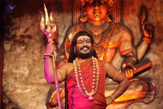 A smiling man with long dark hair and a full beard stands in front of a large orange-lit carved deity murti with many necklaces and multiple arms. He holds a gold trident in his raised right hand, wears a rust-orange wrap with gold trim, layered beaded purple-and-gold rudraksha-style malas, gold armlets and bangles, and a white horizontal tilak on his forehead. His left hand rests on his hip. Swamiji (probable): long swept-back hair, ornate orange clothing, and ceremonial beaded necklaces match the page's reference to a Swamiji appearance, placing him in the front of the frame as the central figure.
