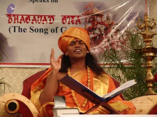 A man seated on a cushioned chair speaks while gesturing with his right hand raised, a discourse setting. He wears an orange turban with a small red bindi, a saffron-orange garment with gold border, and a gold ceremonial necklace of beads, with long dark hair visible at his shoulders. An open blue-covered book rests on a wooden stand before him. Behind him a banner reads Bhagavad Gita (The Song of God) and shows a red chariot scene with horses, beside a tall golden oil lamp and green potted plants. Swamiji (probable): long swept hair, orange turban and ornate gold necklace, seated as the discourse speaker.
