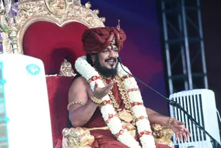 A man seated on an ornate gilded throne with a red cushioned back, speaking into a microphone on a stand. He wears a maroon-red turban with a small gold ornament, a red-brown sannyasi robe, multiple white jasmine garlands with red flowers, a heavy gold necklace, and gold bangles. He has a full dark beard and gestures with his right hand open, mid-discourse. A white cooler unit and stage truss are visible at the sides. Probable Swamiji's cues are the swept turban, the ceremonial gold ornaments and the maroon-red robe, seated in the throne role of a discourse. Only one person is physically present.