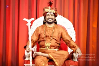 A photograph of a smiling man seated cross-legged on an ornate white and red throne-style chair against a red draped curtain backdrop. He wears a saffron-orange kurta, a matching wrap, a layered rudraksha mala and a large gold necklace, with gold earrings and a saffron-and-maroon turban topped with a gold ornament and red plume. He holds a white staff in his left hand and rests his right hand on the armrest.