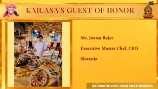 A gold-and-red graphic slide titled "KAILASA'S GUEST OF HONOR" with a small painted portrait of a smiling figure in orange-saffron head wrap at upper left, and a sacred emblem at upper right. A framed photograph shows a woman in a white chef's jacket standing behind a buffet table laden with fruit platters, grapes, sliced vegetables, tomato and mozzarella arrangements, pastries and orange juice glasses, in a stone-walled restaurant with paintings and red chairs. The slide's right side lists a named guest with her title and country. Footer reads Navaratri 2022 | 42nd Chaturmasya.
