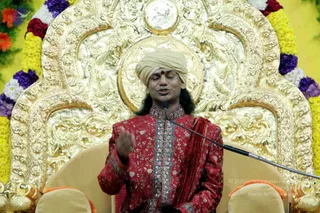 A man seated on an ornate gold throne with carved floral and scrolled panels, framed by garlands of yellow marigolds and purple and white flowers. He wears a cream turban with a red bindi, a red embroidered sherwani with silver and blue trim, and a red shawl over one shoulder. He speaks into a microphone on a stand, one hand raised in gesture, eyes closed. a person: a person in a turban and red embroidered sherwani seated on a golden throne with long dark hair visible behind the shoulders, matching the page's a person reference.
