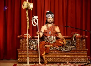A man seated cross-legged on an ornately carved wooden throne upholstered in leopard-print fabric, in front of red stage drapery. He wears an orange-red dhoti and upper cloth, a leopard-print headband with a crescent emblem, multiple rudraksha malas, and gold ornaments and bangles; he holds a white ornamental staff topped with a carved figure and a leopard-print drum above him. A microphone stands at his right. Swamiji (probable): long hair and saffron-orange ceremonial dress with rudraksha malas and a crescent crown mark a candidate in the central seat.