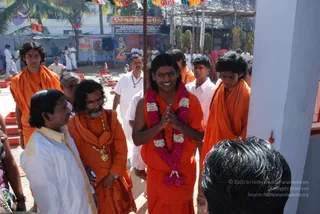 Outdoor temple-festival scene with a central young man in saffron robes and a pink-and-white flower garland, hands pressed together in greeting with a small red tilak on his forehead. Around him, saffron-clad attendants stand with bare-chested and white-clad devotees; an older man in a white shirt and a bearded man in orange robes with a gold-chain pendant stand to the left. Behind them, a canopy of orange and green flags hangs over a stall with a painted deity panel and a red-and-yellow shrine, with a white-clad crowd and red seating blocks in the courtyard. Roughly five to six people are visible.