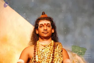 A bare-chested man with long, swept-back dark hair stands before a painted blue-and-gold backdrop, wearing a leopard-print wrap draped over one shoulder. He has a tall topknot adorned with a rudraksha band, white horizontal tilak lines on the forehead with a red bindi, and multiple rudraksha malas and ceremonial gold beaded necklaces across the chest. A white band is wrapped around one wrist.