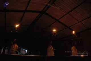 Photograph of three men in plain white dhotis standing in a dimly lit hall with a corrugated metal roof and exposed dark beams. Each raises an oil lamp overhead, with flames visible on the lamps. The man on the left wears a beaded necklace and the one in the centre wears a cord and beads; the man on the right is partly in shadow. A row of framed photographs hangs on the wall in the background, and a small flame glows at the lower right. The scene suggests a lamp-waving ritual such as aarti.