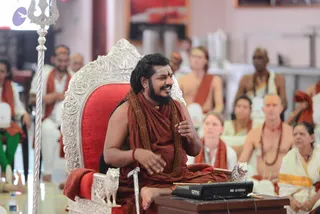 A bearded man with long dark hair sits on an ornate silver-and-red throne with carved lion armrests, wearing a rust-orange wrap, a long rudraksha mala and a gold necklace, with a vertical sandalwood mark on his forehead. He smiles and gestures with his right hand while speaking; a silver trident stands to the left and a small black audio mixer sits on a table in front of him. Seated devotees in orange and white clothing, including a woman in a white sari at right and people wearing orange shawls, fill the hall behind him. The cues are distinctive: long swept hair, saffron-orange clothing, rudraksha mala and gold necklace on a throne, so this is an appearance-based a person in person.