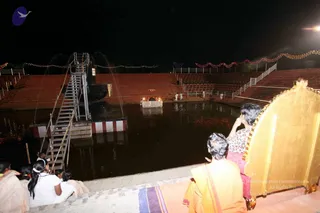 Night photograph of an outdoor temple tank at a KAILASA ashram, with dark water, a metal ladder on the left and terraced red-tiled steps rising behind. Strings of coloured lights line the upper edges. In the centre, a small white-draped platform on the water holds lit oil lamps and offerings, with a few white-clad attendants standing near it. In the right foreground, a person in a saffron-orange garment sits on the steps beside a gold-trimmed seat, with another seated person in a floral top and pink cloth nearby. On the left, several people in white garments and a white-clad woman with a white hair band sit on the steps watching the ceremony.