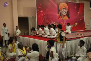 Indoor stage event with a red-carpeted platform and a large red backdrop showing a smiling man in an orange turban and saffron garment beside the label "Anandotsava 2010". The backdrop depicts a portrait; a man in white with a dark neck garland stands at left holding a microphone, and a woman in white with a dark garland stands beside him. Several children and young people in white dhoti-style or white kurta outfits sit and stand on the stage, one boy in a blue vest with gold trim and white dhoti stands front right. Seated audience members in white and yellow shawls, including a woman in a yellow shawl, watch from floor mats at the lower left and right foreground.