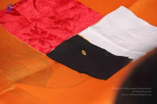 Close-up photograph of an orange silk cloth with a rectangular patchwork of red velvet with embossed floral pattern, plain white fabric, and black fabric. A small yellow faceted gem sits on the black patch. A blurred dark object is at the lower right edge. No people are visible.