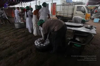 Outdoor food preparation area under a blue tarp canopy on bare, muddy ground. Several volunteers in white clothes and green hairnets work at steel tables and stand near large metal cooking pots. A man in a brown shirt and dark trousers bends over a pot lined with aluminium foil, while a woman in a brown sari stands nearby. A white pickup truck is parked behind the tent beside a tent wall. Steel plates and bowls sit on the table.