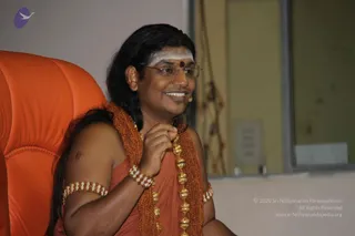 A photograph of a smiling man seated in an orange leather chair, speaking into a small headset microphone. He has long dark hair swept back, a white vertical tilak with a red dot on his forehead, and wears thin-framed glasses. He is draped in orange cloth and wears orange beaded necklaces with gold bead accents, plus beaded bracelets on his wrist and upper arm. His right hand rests near his chest. Behind him are a pale yellow-green wall and a window. The appearance is consistent with the a person cues of long hair, orange clothing and ornate beads, but identity is uncertain from appearance alone.