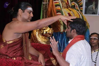 A seated man in a maroon-red robe with gold border and a gold-and-brown beaded necklace places his right hand on the head of a man in a white shirt with a red shawl, performing a blessing. Behind them stands a large golden lion throne and a gold-and-blue decorated altar draped with garlands of yellow, orange and red flowers. A third man in white with a white forehead mark and beaded necklace looks on at right. The seated figure has a shaved-style hairline with a white tilak and his arm bears a scar.
