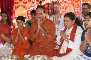 Devotees sit in a tented ritual space with red and gold draped fabric behind them, offering prayers with palms pressed together in anjali mudra. A man in an orange kurta with a rudraksha-style necklace sits at centre with eyes closed, next to an older woman in a white sari with a red shawl and a young girl in orange robes with a beaded mala. Behind them a bald man in white with a red-and-white forehead mark and a beaded necklace also prays. Silver plates with flowers and offerings sit in the foreground, and several more people in white and red clothing are seated at the edges. Seven people are visible, all participating in a puja or satsang setting.