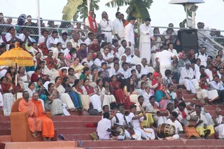 Outdoor ceremony on tiered red-brick steps before a large crowd of seated devotees, many in white with red or saffron shawls. At lower left, two saffron-robed figures sit on an ornate orange throne-style chair under a yellow fringed parasol; one is bald and one wears a saffron wrap. Musicians in white clothes play drums, a horn and a wind instrument at lower centre. Standing figures in white and a man in white with a red shawl stand along the upper steps behind a chain-link fence, with palm fronds visible at top.