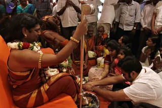 A seated figure with long dark hair, wearing an orange-and-gold saffron garment, layered red, white and green flower garlands, a rudraksha mala and gold bangles, extends a hand toward a tall silver ritual stand while sitting on an orange cushion. a person, in appearance-based identification with long swept hair, saffron-gold clothing, rudraksha mala and ceremonial gold ornaments. Before the figure, a man in a white shirt kneels and holds a brass plate with offerings, and seated devotees including a young girl in pink and a woman in a red sari with a child wait nearby. Several shaven-headed sannyasis in orange robes sit behind, and a crowd of standing men in white shirts watches. This is a padapuja or pada puja foot-worship ritual in a hall with orange seating.