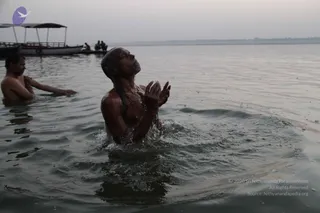 A photograph of a river scene at dawn or dusk, with hazy grey sky and a flat boat with a canopy in the background. In the foreground, a bare-chested man stands waist-deep in the river, head tilted back, eyes lifted, palms pressed together in prayer as water splashes around him. To the left, another bare-shouldered man stands partly submerged, hand raised near the water. Two more figures sit on a distant boat. The scene suggests a ritual bathing or prayer in the Ganga at Varanasi.