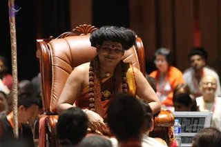 A person with swept-back dark hair and a red bindi sits smiling in an ornate brown leather armchair on a stage, wearing an orange-and-saffron garment with a gold-toned shawl, multiple rudraksha malas and a gold pendant necklace. a person is seated centrally as the focal figure, with a seated audience of people in orange and white lanyards in the foreground and background. A water bottle and a laptop screen are visible at right, and a metal pole stands at left.