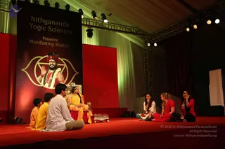 Stage photo of a Nithyananda Yogic Sciences 'Manifesting Shaktis' session. A large banner reading 'Nithyananda Yogic Sciences Presents Manifesting Shaktis' shows a painted figure in a red turban and white flower garland, raised hand, beside a gold yantra-like emblem. In the centre, a woman in a yellow sari sits on an ornate gold throne with a red cushion. Two boys in yellow and orange clothing sit on the left, a young man in white kurta sits on the red stage floor, and two women in red attire, one holding a microphone, sit on the right. Red backdrops, stage lights and a brass tray with flowers complete the scene.