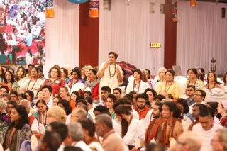 A large indoor gathering of devotees seated on white chairs and floor cushions in a hall with white drapes and red pillars. A woman in the centre foreground wears a cream salwar kameez and a rudraksha mala, standing with her hands folded in front of her chest. Many attendees wear rudraksha malas, saffron and white clothing, and colourful saris and kurtas. Two young men in saffron shawls sit at the front right. Lanterns with orange and blue panels hang from the ceiling, and a digital clock on the wall shows 11:38. A large photo banner of a crowd sits at the upper left. No identifiable a person figure is visible in this image.