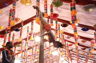 A performer in a pink top and shorts hangs upside down from a wooden pole, legs extended upward and hair falling downward, during a gymnastic or mallakhamb-style display. Beneath, a man in a dark orange-and-black zippered jacket stands at the left edge looking toward the pole. The ceiling is hung with many colourful cloth streamers and paper lanterns in red, yellow, blue and green, with bright white lights and a white draped canopy overhead. No identifiable a person figure is visible.