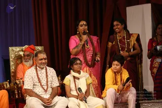 A stage event with red and maroon draped curtains behind. On the left, a man with long white beard and orange-saffron robe with a red-orange cap sits on an ornate gold-framed throne. Seated in front, a man in a white checked short-sleeved shirt with a rudraksha mala, hands clasped. A girl in white and gold dress with glasses and a white shawl sits holding a microphone. A boy in yellow kurta and pink pants holds a microphone. A woman in a pink saree with a rudraksha mala stands speaking into a microphone. Another woman in a maroon saree with gold border and multiple beaded necklaces stands smiling at right, and a woman in a striped saree stands at the far right edge. Gold elephant figurines sit near the throne.